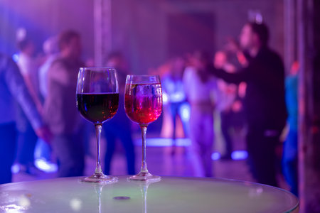 Two wine glasses on a table at a lively party with colorful lights and people dancing in the background.の写真素材