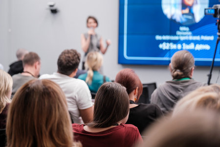 Diverse group sitting in audience listening to a speaker at professional conference or seminar. Blurred presenter and screen in background.の写真素材