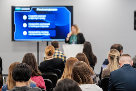 Business speaker presents to audience at conference with large screen display. Participants attentively listen and take notes in modern work environment.の写真素材