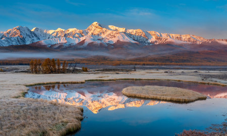 Sunrise highlights snowcapped mountain peaks, reflected in calm water, surrounded by autumnal grassland. Captivating natural scene featuring vibrant lighting and serene atmosphere.の写真素材