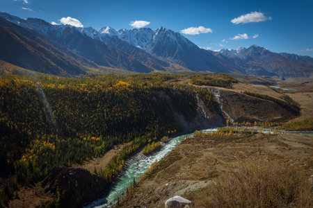 A stunning mountain river flows through a forest, surrounded by snowy peaks and valleysの写真素材
