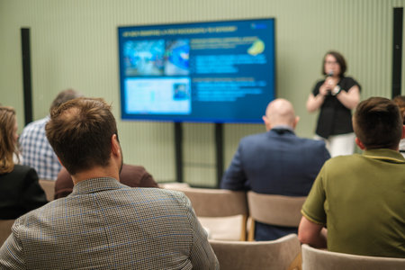 Audience listening to a lecture with speaker, focusing on a screen displaying information.