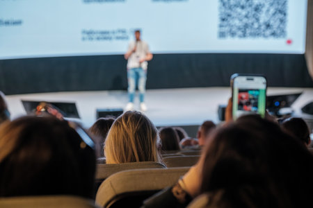 Conference setting with speaker presenting on stage, viewed from audience perspective. Smartphone capturing the eventの写真素材