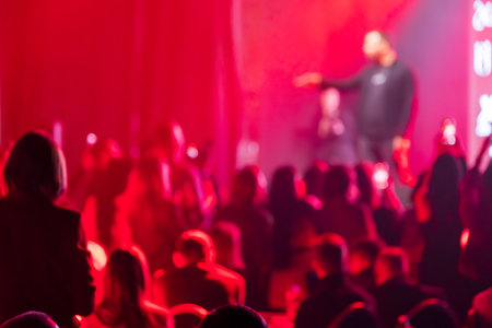 Blurry red-lit audience observing a presenter or performer on stage during an event.の写真素材