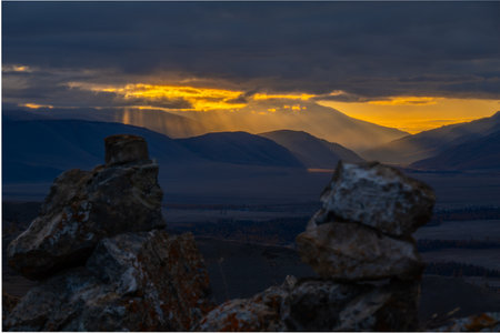Warm sunset breaks through clouds illuminating distant mountains while rugged rocks in foreground...