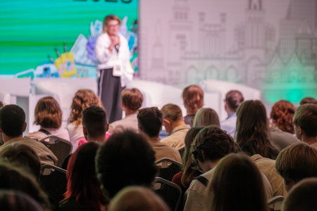 Audience fills seats as presenter speaks on stage with colorful backdrop bright lights creating energetic conference atmosphereの写真素材