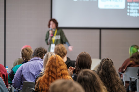 Presenter in green shirt speaks into microphone before attentive crowd in conference room. Bright screen displays slides as participants listen, learn, and take notes during group education sessionの写真素材