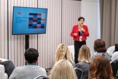 Speaker in red blazer presents to attentive audience during conference session with colorful monitor...