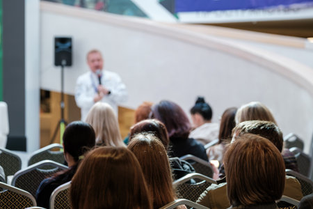 Crowd of attendees listens to speaker delivering talk at modern venue. Audience sits in rows, stage behind, equipment and banners visible, event atmosphere focused and professional.の写真素材