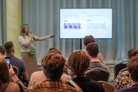 Presenter speaks into microphone while pointing to projected slide as attendees watch. Bright room, conference setting, engaged audience, learning vibe, professional presentation, sharing information.の写真素材