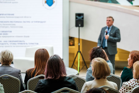Audience attends presentation in conference hall while speaker delivers talk with microphone and large screenの写真素材