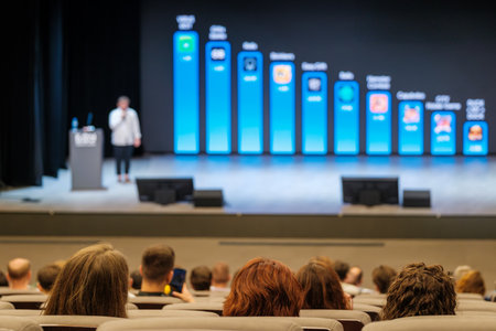 Audience watches speaker on stage with blue bar chart backdrop during conference