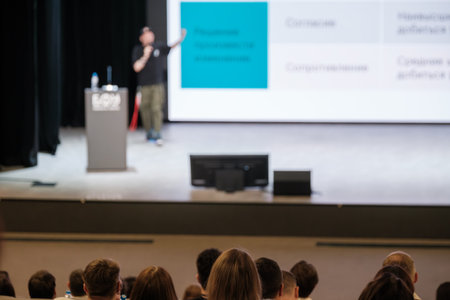 Speaker presents at conference stage with podium, screens display charts, audience seated listening attentively, microphones and water bottle on podium, professional event atmosphereの写真素材