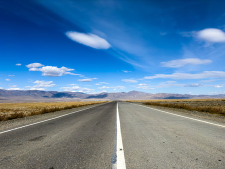 Desert highway stretches to horizon under clear blue sky with scattered clouds distant mountains open landscape evokes freedom travel and adventureの写真素材