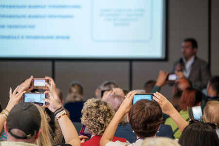 crowd of attendees watch speaker deliver presentation many capture moments on smartphones creating...