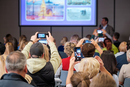 Crowd watches speaker on stage at conference while attendees record slides with smartphones and cameras creating dynamic social video momentの写真素材