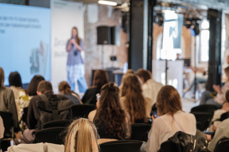Conference hall with speaker on stage delivering presentation to attentive audience, attendees sit...