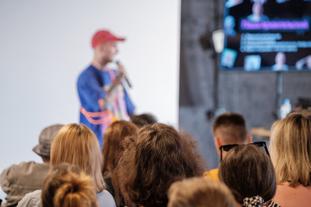 Crowd listens to presenter on stage during conference with large screen displayの写真素材