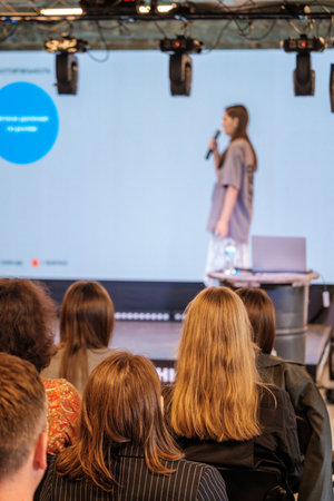 Female presenter on stage speaks into microphone before audience at conference with blue screen background and laptop on podiumの写真素材