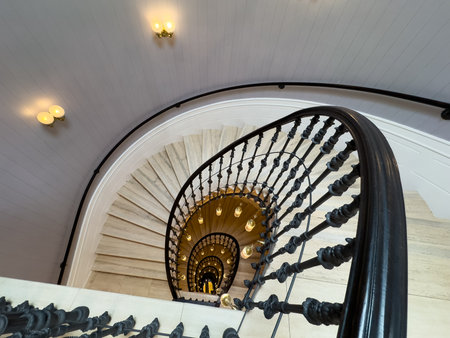 Spiral staircase shows ornate black balusters marble steps curved railing and warm light fixtures creating dramatic downward perspective and elegant historic atmosphereの写真素材