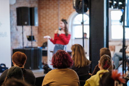 Female presenter in red sweater speaks into microphone guiding audience during workshop in brick venue with stage lights creating vibrant modern atmosphereの写真素材