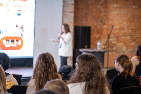 Presenter speaks to audience at conference in brick space. Attendees listen as large screen shows content and microphone stands nearbyの写真素材
