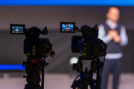 Two cameras on tripods film a speaker on stage with blurred audience and blue lighting.の写真素材