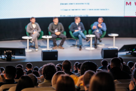 Panel of four speakers on stage during live conference, green chairs, round tables, audience listening attentively, professional eventの写真素材
