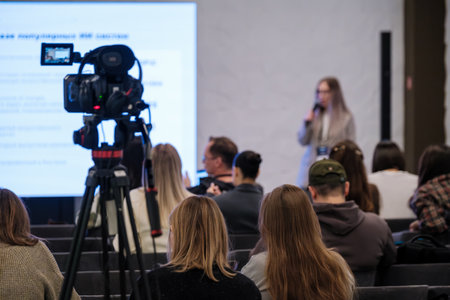 Audience watches presenter at seminar as camera captures scene on tripod in bright modern room