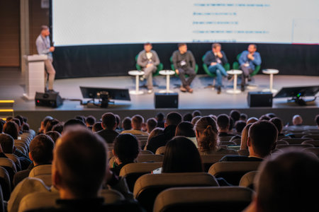 Audience watches panelists on stage during conference, panelists seated with small tables, large screen behind, stage lighting creates formal atmosphereの写真素材