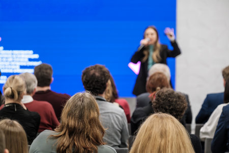 Presenter speaks to attentive crowd during corporate conference. Bright blue backdrop highlights...