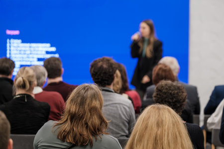 Audience focuses on speaker during conference against bright blue screen. Attendees sit in modern...