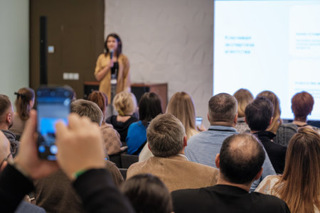 A speaker presents to a crowded conference room while attendees watch large screens and capture moments with mobile devices. The scene conveys learning, engagement, and professional gathering.の写真素材