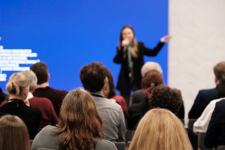 Speaker delivers presentation to attentive crowd in conference room. Blue backdrop creates modern setting. Audience listens closely feels curious inspired focused during talk.の写真素材