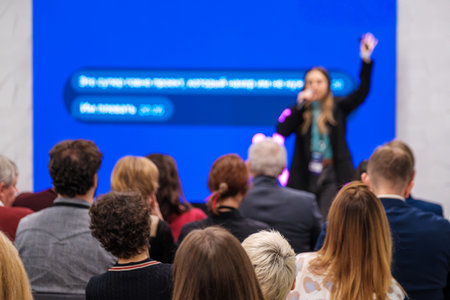 Speaker on stage addresses crowd at conference while audience listens. Bright blue backdrop...