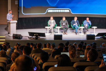 Panel discussion on stage featuring five speakers in green chairs, audience in auditorium watches...