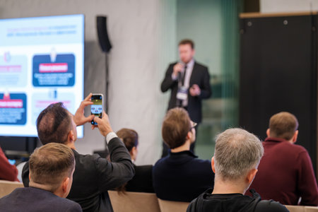 Business conference scene with presenter on stage and engaged audience. Attendee films talk on mobile phone. Modern venue with bright screens in background.の写真素材
