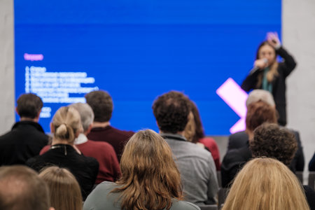 Speaker addresses audience at conference with large blue screen backdrop. Attentive crowd listens...