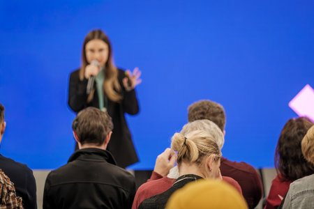 Audience listens to speaker during workshop. Presenter uses microphone and gestures as blue background sets stage atmosphere. Attentive crowd wearing casual clothing.の写真素材