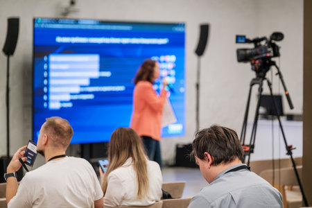Conference room scene with speaker on stage delivering content to audience while camera records. Large blue screen shows charts. Attendees watch smartphones and take notes.の写真素材