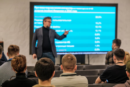 Speaker presents data to attentive audience modern conference room. Blue screen displays statistics...