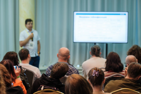 Presenter speaks at conference to attentive audience during seminar. Screen displays slides.