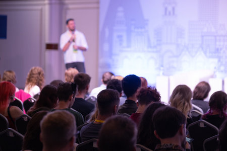 audience sits in conference hall listening to speaker on stage, bright backdrop cityscape displayed,...