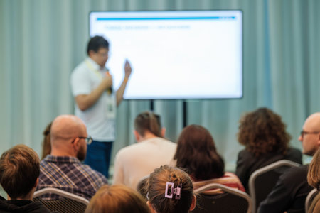 Presenter speaks to seated audience in conference room with large screen monitor. Attentive...