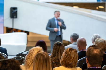 Presenter in suit speaks to seated crowd at conference. Audience listens attentively in bright...