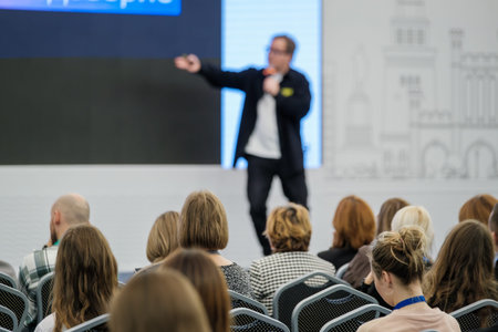 Audience seated listening to presenter on stage during business conference. Focus on engagement,...