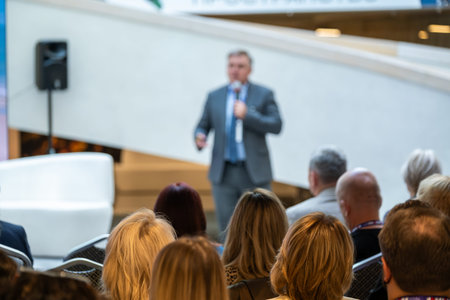 Man in suit delivers talk to attentive audience in bright modern venue. Presenter holds microphone...
