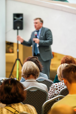 Professional speaker addressing seated audience during business conference. Attendees listening and...