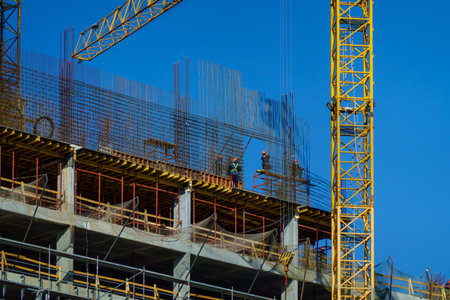 Construction workers installing steel rebar on high rise concrete building, yellow crane against...