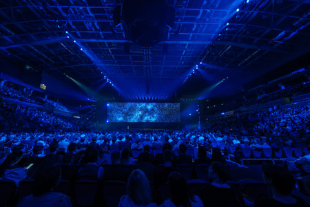 Packed conference auditorium bathed in blue light, audience watching large LED screen showing starry galaxy visuals, immersive stage production, live event atmosphere.